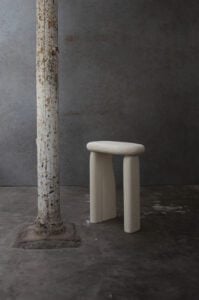 Windows of Bo Bardi stool in butter-colored material with a rounded seat on solid slab legs beside a raw concrete column by Linde Freya Tangelder