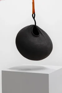 Movement Phase 2 sculptural vessel in matte black stoneware with soft folds, suspended by an orange strap above a white plinth, by Catalin Filip