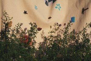 Cream woven blanket with scattered embroidered motifs in teal, brown and red, photographed behind wild flowering plants in sunlight