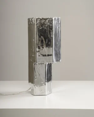 Lost Table Lamp in polished aluminum on a table, angled view.