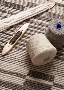 Wooden weaving shuttle and pick-up stick resting on a striped flatweave textile, with two spools of undyed natural yarn