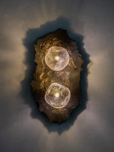 Asea No. 25 wall sconce with two glowing textured hand-blown glass globes set in a dark wavy stoneware plate, by Ceren Gürkan