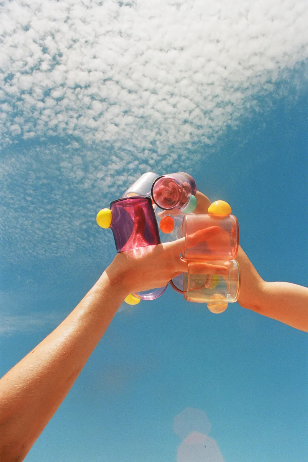 Two hands toasting with Bubble Cups in hand-blown glass, one filled with a pink drink, against a blue sky with clouds, by Sticky Glass