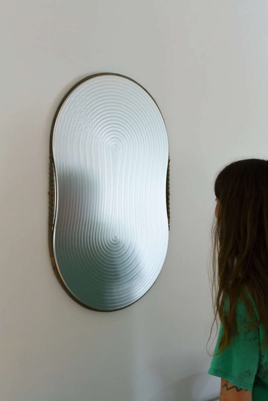 Oval wall mirror with a rippled, textured glass surface in a thin metal frame, mounted on a pale wall