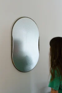 Oval wall mirror with a rippled, textured glass surface in a thin metal frame, mounted on a pale wall