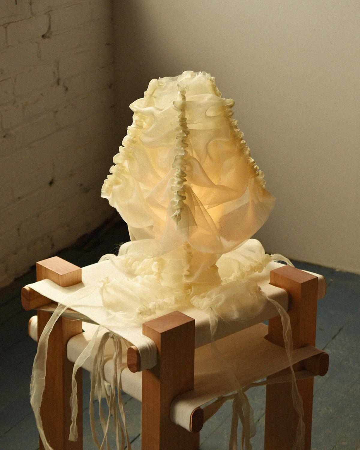 Chloé table lamp in a pale, gathered reflective textile veil glowing softly on a wooden stool, by Atelier Alz