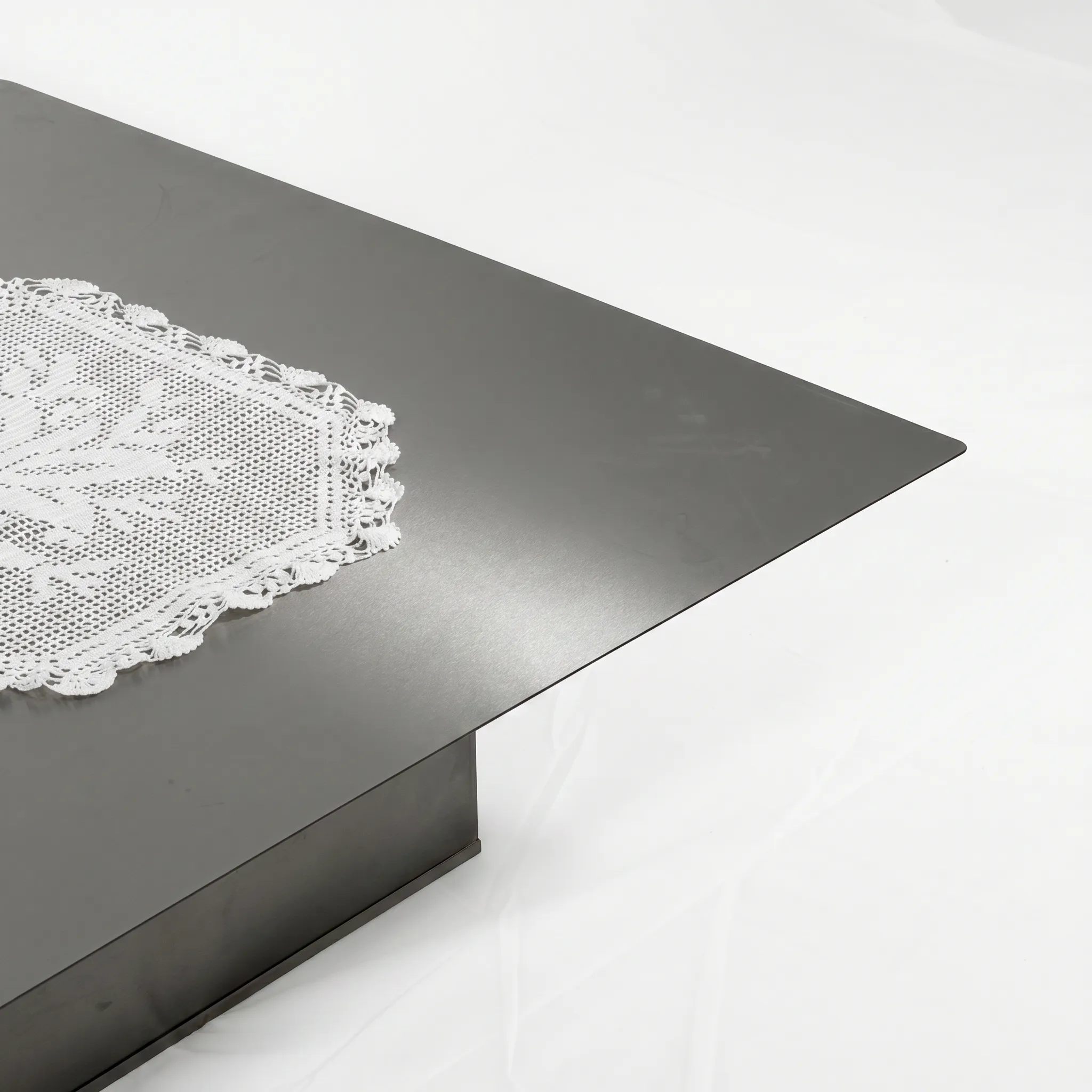 Low square coffee table in dark stainless steel with a thin overhanging top on a block base, a white lace doily laid on its corner, by OFAA