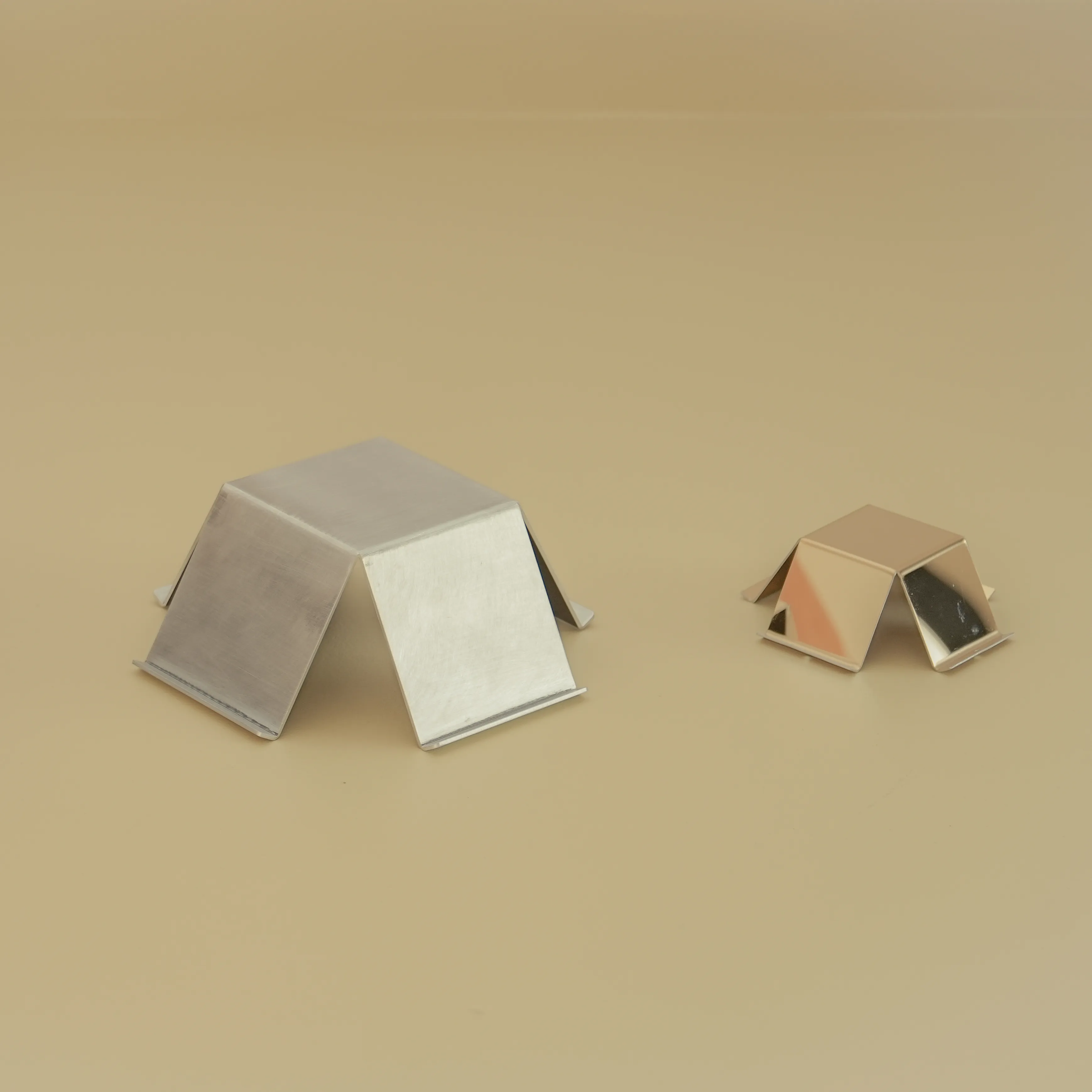 In By On Object 01 in brushed aluminum and Object 02 in gold mirror-polished stainless steel, two folded geometric forms on a beige surface, by seamlessnote