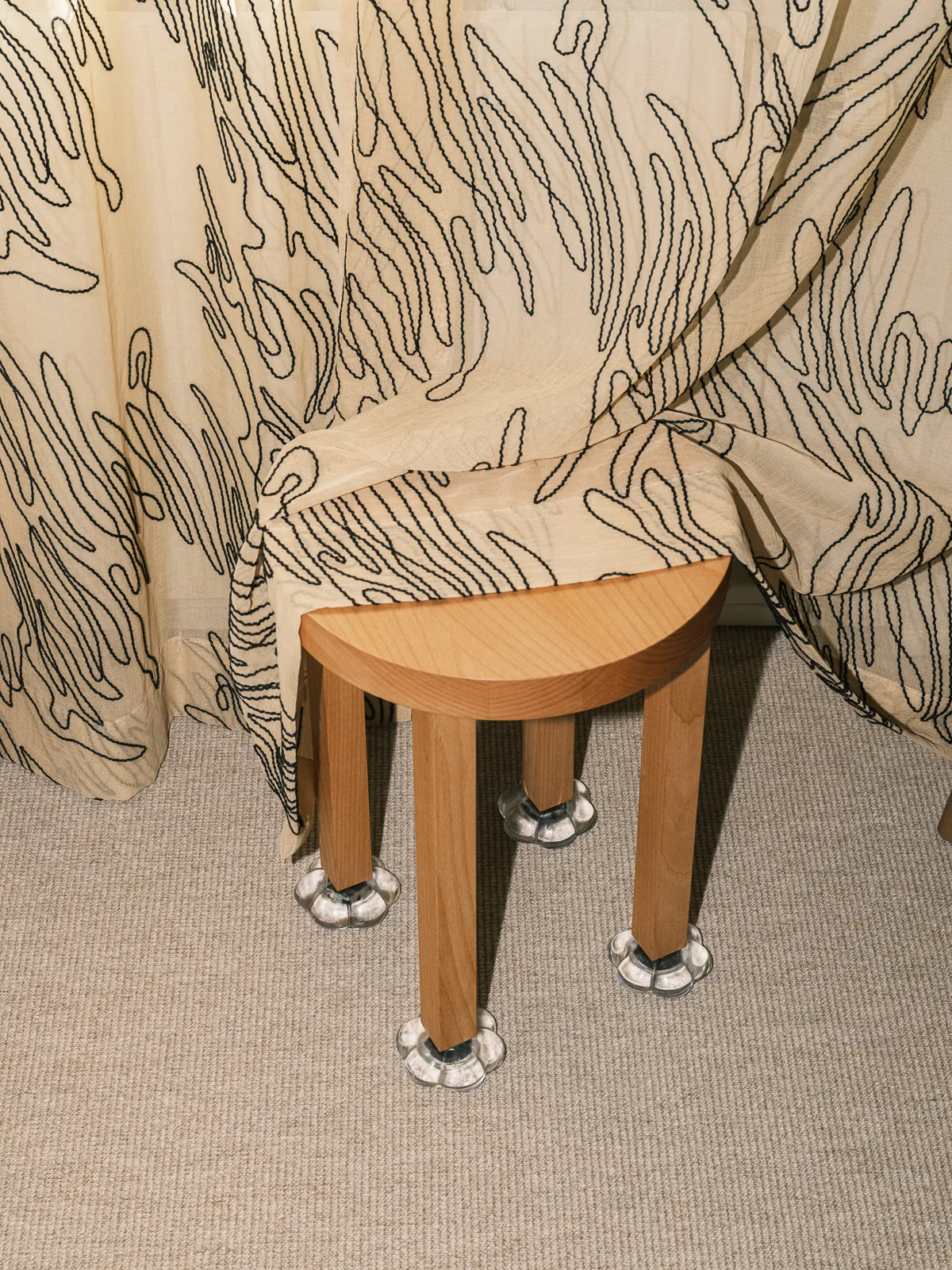 New Shoes stool with a thick half-round alder wood seat on four legs, each standing in a small sand-cast glass shoe, in front of a patterned curtain, by Studio Ebba Lindgren
