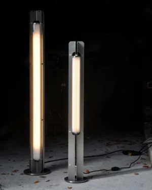 Pill600-70 Floor Lamp in gray stainless steel with a glowing tube light on a floor, front view.
