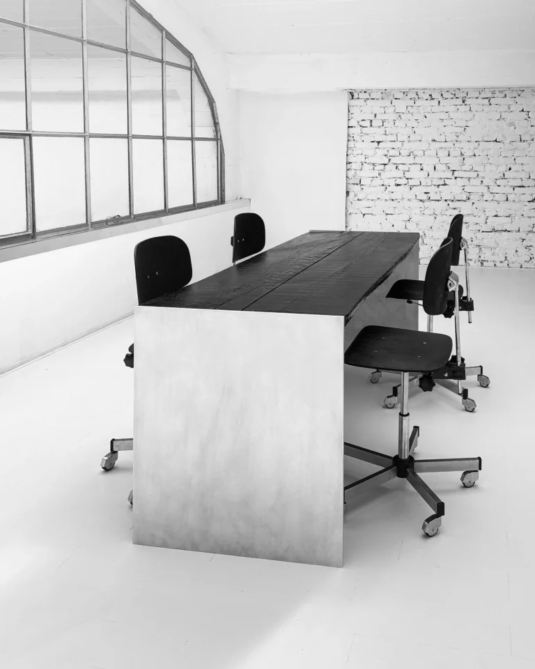 Yakisugi Desk by case studios in a bright studio, black swivel chairs, an arched window and a white brick wall behind