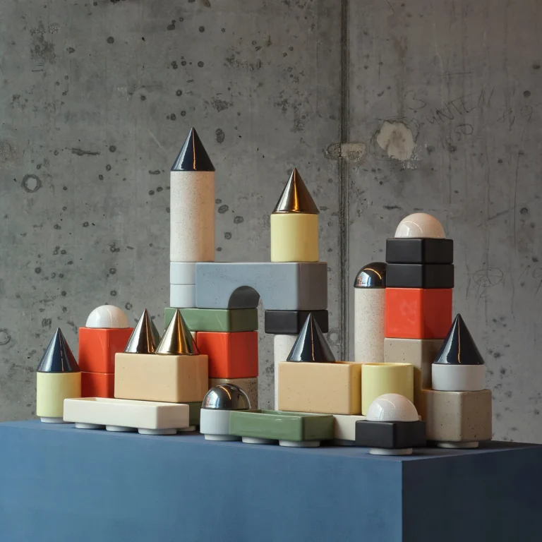 Knstrkt Set Of 14 by Studio Berg, a dense group of colorful stacked blocks and cones on a blue plinth by a concrete wall