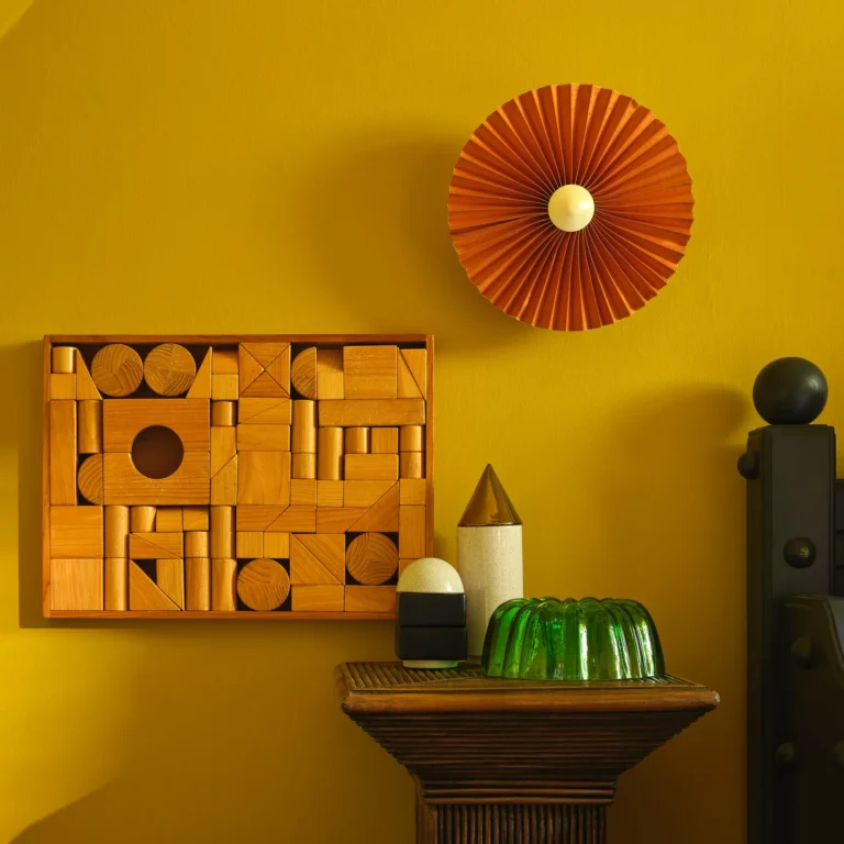 Knstrkt Set Of 14 by Studio Berg on a wooden pedestal against a yellow wall, near a wall of wooden toy blocks