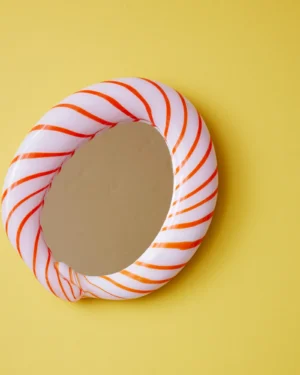 Red Candy Mirror by Studio Berg, round wall mirror in twisted glass with orange-red stripes, hung on a yellow wall
