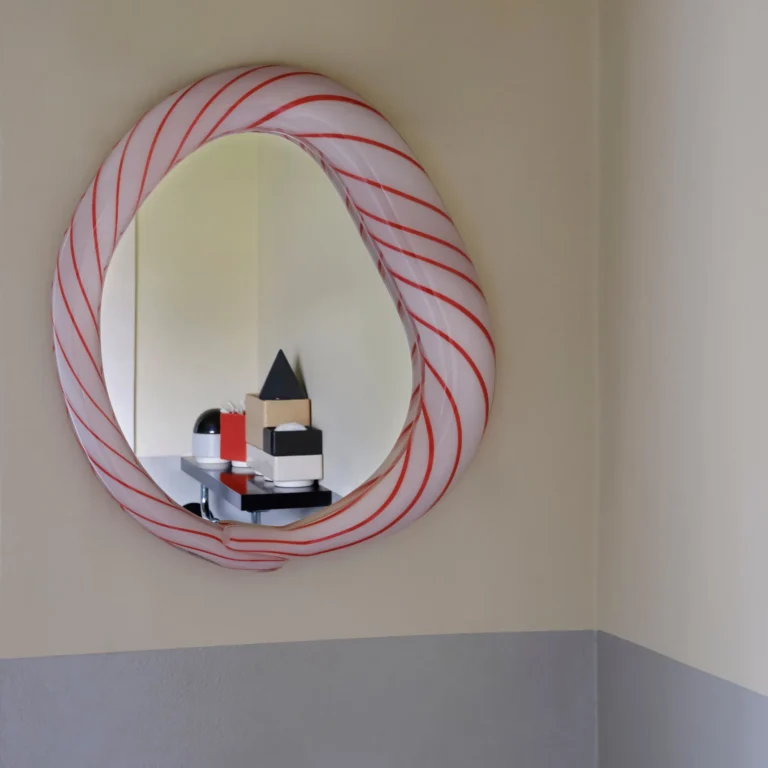 Red Candy Mirror by Studio Berg in a hallway corner, reflecting a shelf with small geometric objects