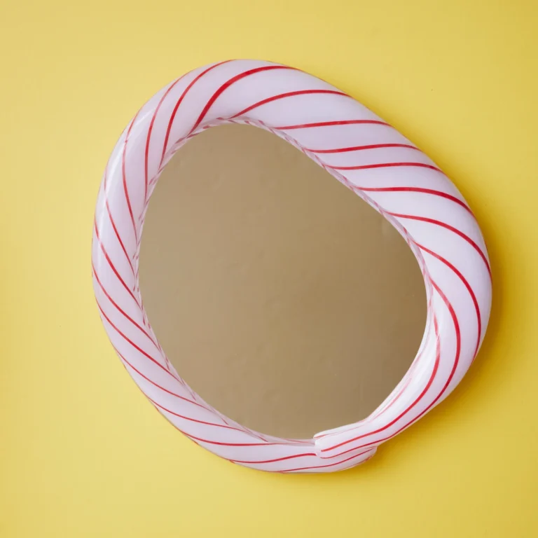 Red Candy Mirror by Studio Berg seen from a slight angle, showing the glass frame's overlapping twisted end on a yellow wall