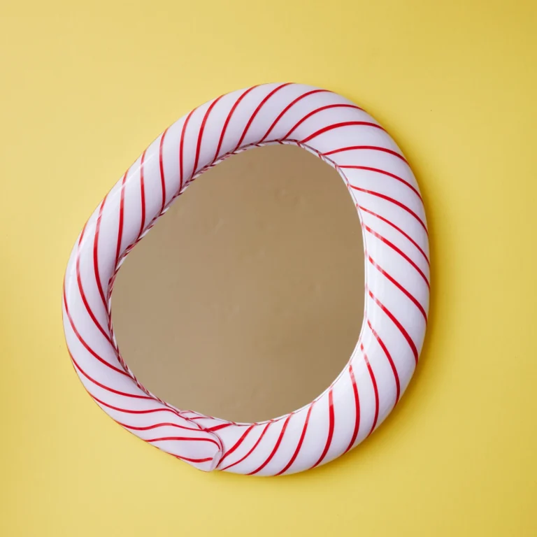 Red Candy Mirror by Studio Berg, front view of the irregular oval frame with thin red spiral stripes, on a yellow wall