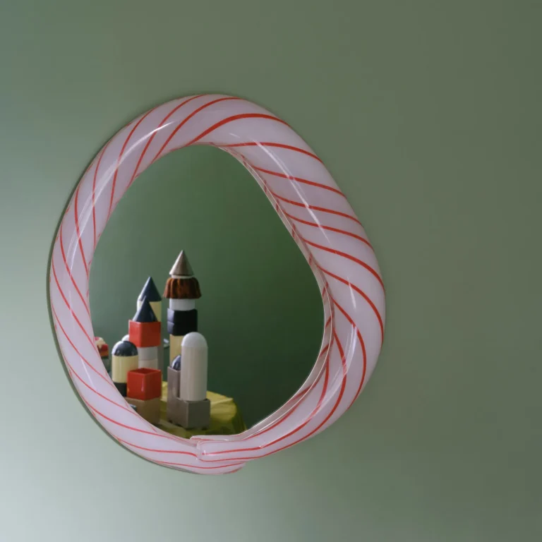Red Candy Mirror by Studio Berg on a green wall, its twisted glass frame reflecting a group of small tower-like objects
