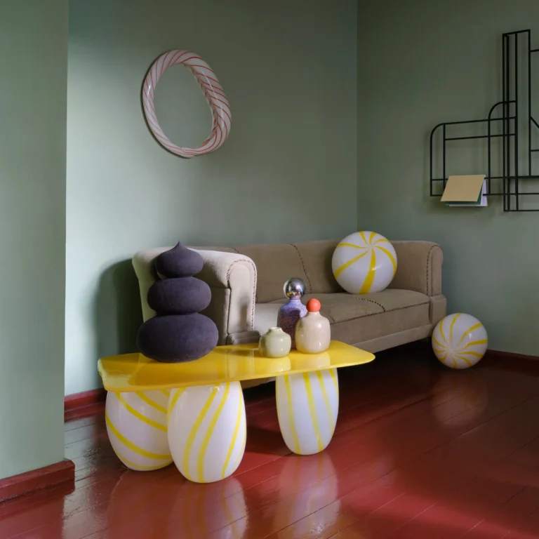 Red Candy Mirror by Studio Berg in a green living room, hung above a sofa and a yellow table with striped glass legs