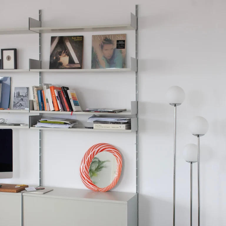 Red Candy Mirror by Studio Berg leaning on a sideboard beneath a shelving system, beside globe floor lamps
