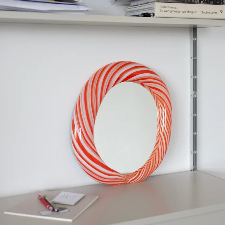Red Candy Mirror by Studio Berg resting on a shelf against a white wall, its red and white striped glass frame seen from the side