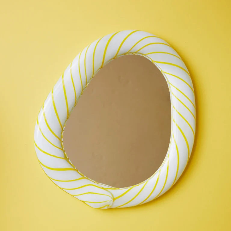 Yellow Candy Mirror by Studio Berg seen from the front, its pebble-shaped mirror and striped glass frame against a yellow backdrop