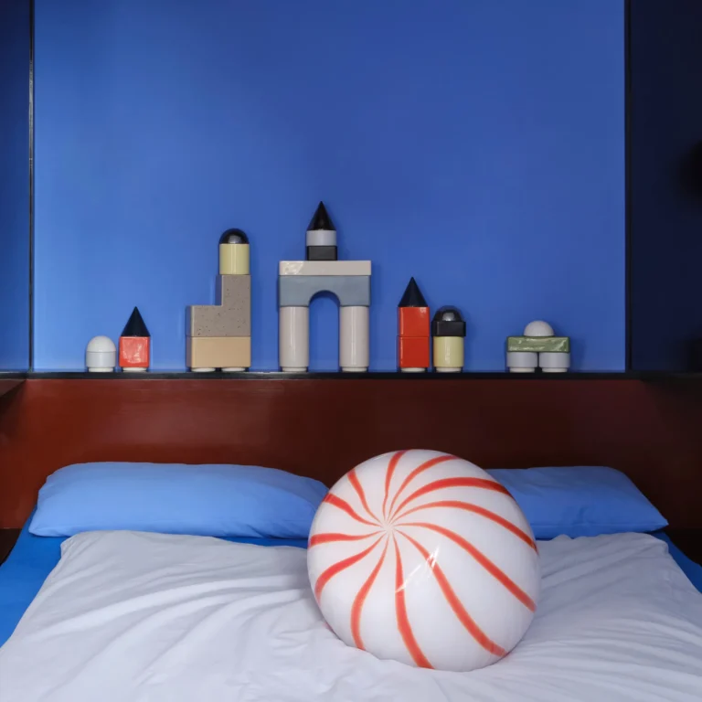 Red Candy Side Table by STUDIO BERG in a bedroom, round striped glass form on a bed below a shelf of small objects