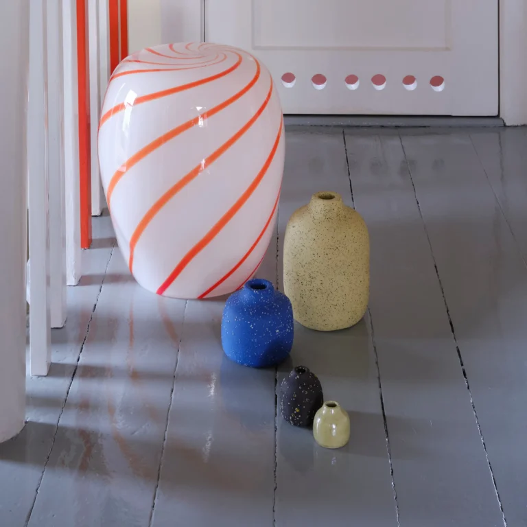 Red Candy Side Table by STUDIO BERG in a hallway, swirled glass piece on a grey floor beside small speckled vases
