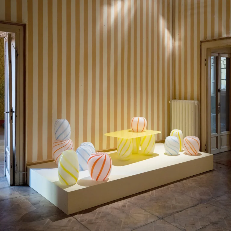 Blue Candy Side Table by STUDIO BERG in a striped-wallpaper room, shown among orange, yellow and blue glass pieces on a low platform