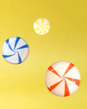 Coatcandy by Studio Berg, three striped glass hooks in blue, yellow and orange-red on white, on a plain yellow ground