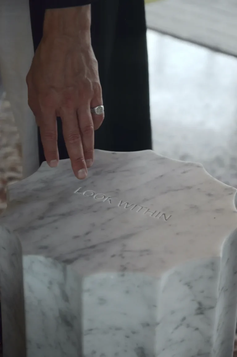 Close-up of the Meditations mirror's marble column, a hand with a ring resting above the engraved LOOK WITHIN