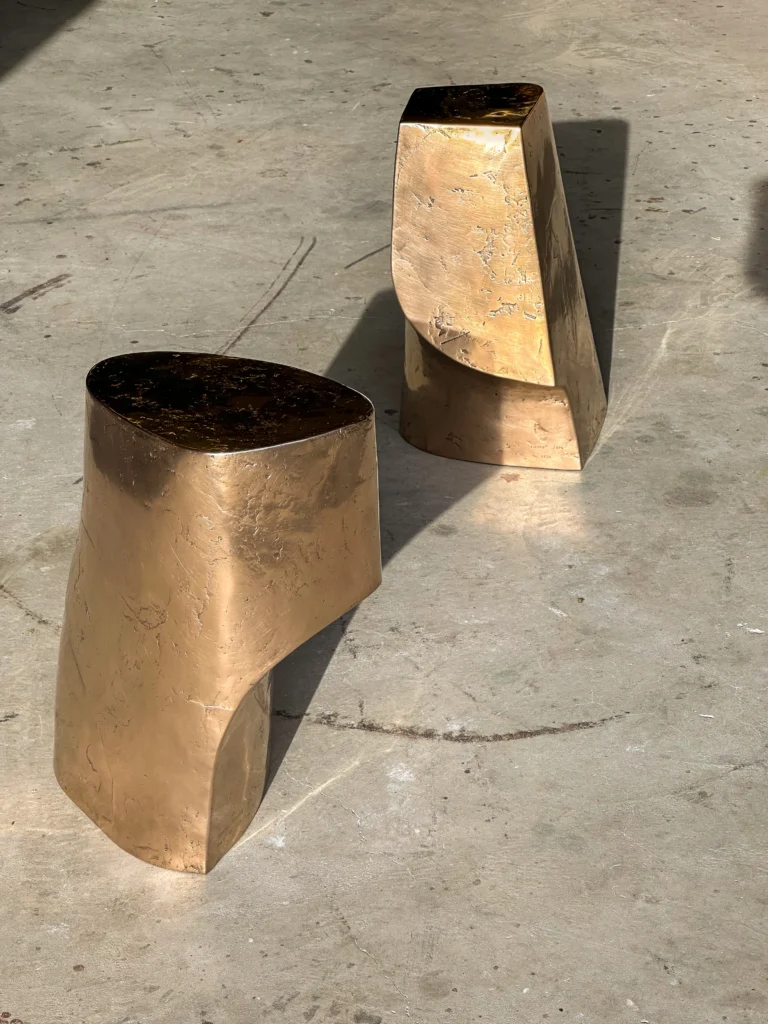 Palm Table I by Vogel Studio on a concrete floor in sunlight, beside a second angular bronze table with a cut-away base