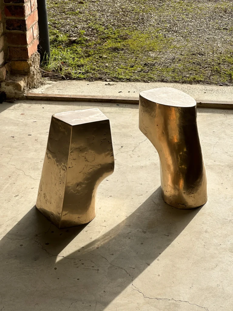 Palm Table I by Vogel Studio in a sunlit doorway, beside a second geometric bronze table, with grass and brick behind