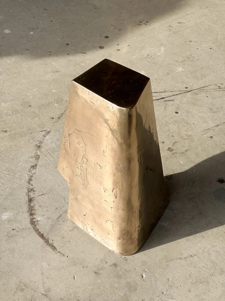Palm Table Il by Vogel Studio seen from the back, a tall tapered bronze form with a dark flat top casting a shadow on concrete