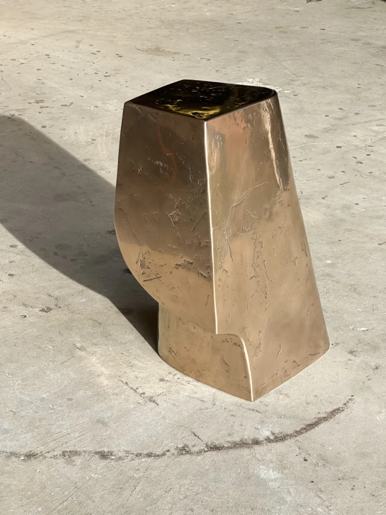 Palm Table Il by Vogel Studio, polished bronze side table with a tapered body and cut-away base, on a concrete floor