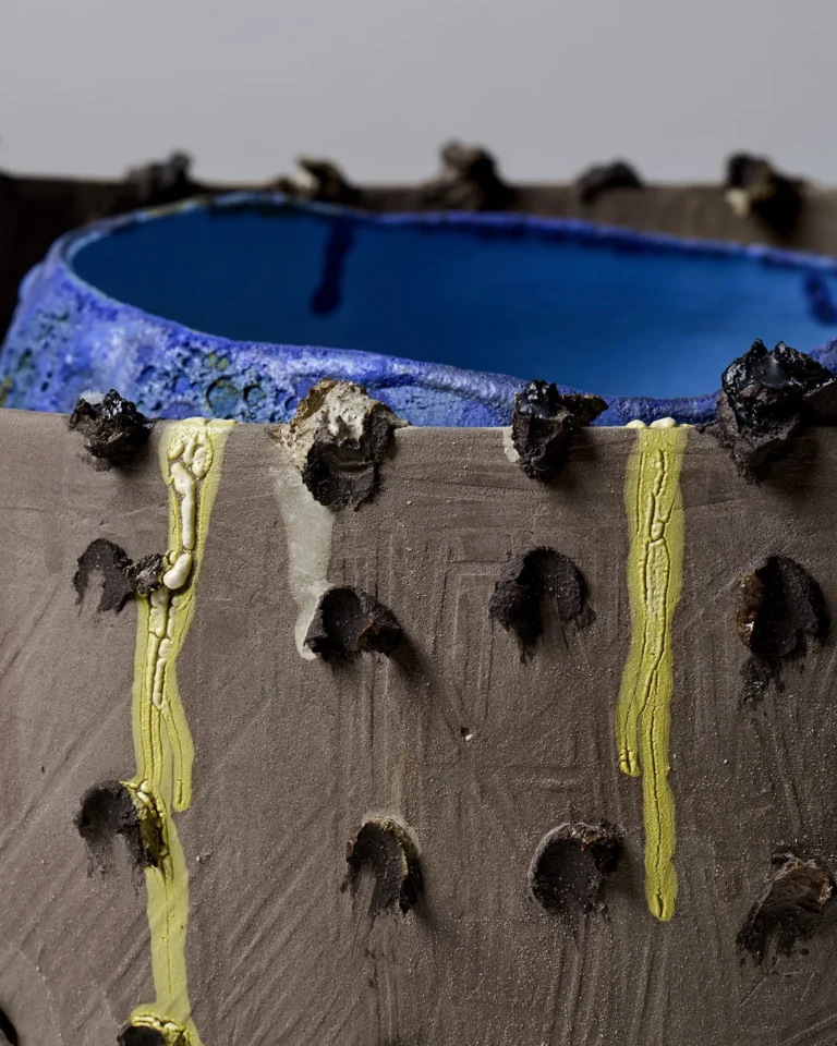 Close-up of the New_love_08 Ceramic Vase's grey wall with yellow glaze drips, pressed dots and blue rim behind